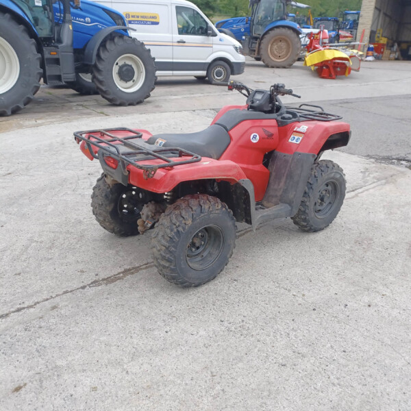 Red Honda ATV parked on a concrete lot with blue tractors and a white service van in the background.