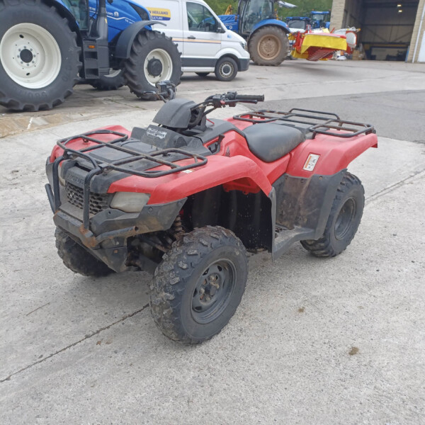 Red ATV in the foreground on a concrete lot, with large blue tractors and a white van visible in the background at an agricultural equipment yard.