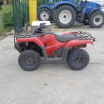 Red Honda ATV in the foreground on a concrete surface, with oversized blue tractors on a trailer and a white van in the background.