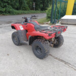 Red Honda ATV quad bike parked on a gravel driveway beside a construction area with a fence and construction equipment in the background.