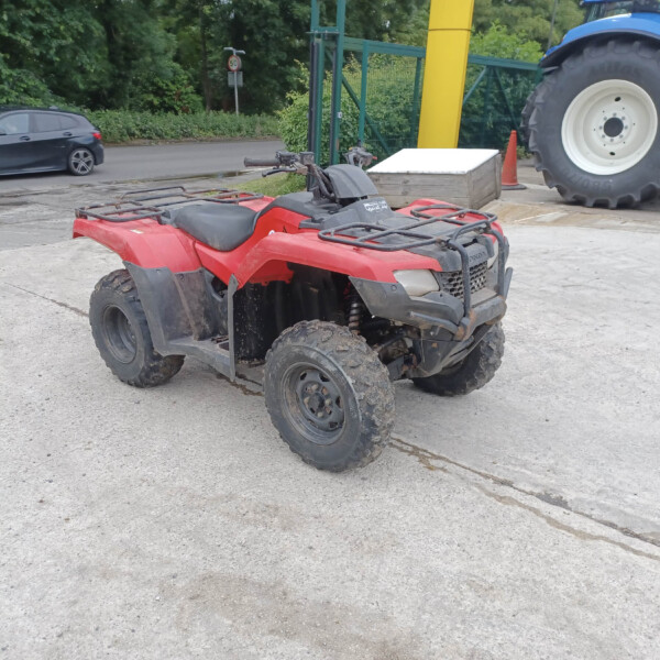 Red ATV parked on a concrete surface with a large blue tractor visible in the background and greenery beyond.