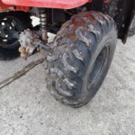 Close-up of a muddy ATV rear wheel, suspension, and a rusted trailer hitch ball in the foreground. The dirt-streaked tire shows off-road treads.