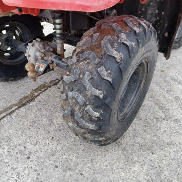 Close-up of a muddy ATV rear wheel, suspension, and a rusted trailer hitch ball in the foreground. The dirt-streaked tire shows off-road treads.
