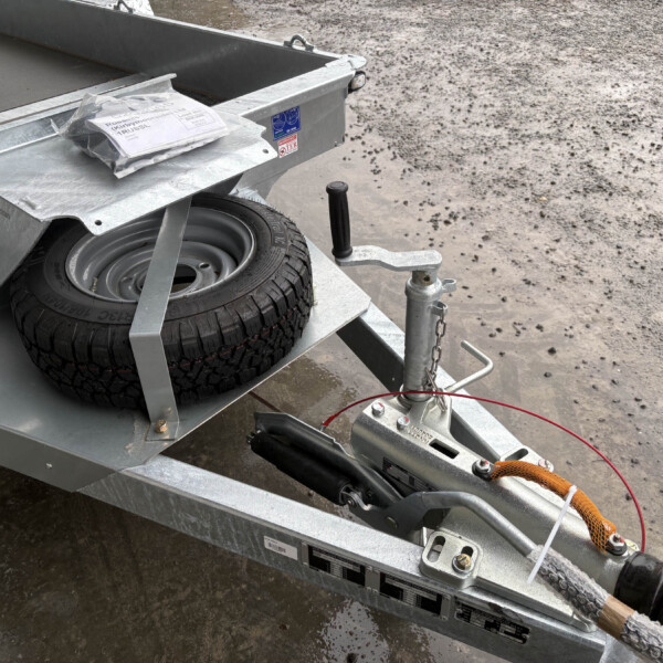 Metal boat trailer with a mounted spare tire, coupling hardware, and a crank jack on a muddy surface.