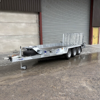 Metal utility trailer with a spare tire parked on wet pavement in front of an industrial building with a large corrugated metal roll-up door and yellow door nearby.