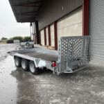 Industrial loading area with a large corrugated metal building, roller shutter door, and a wheeled trailer in the foreground on a wet concrete surface.