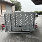Rear view of a gray perforated metal trailer with circular cutouts, parked in a loading yard beside a wooden storage building