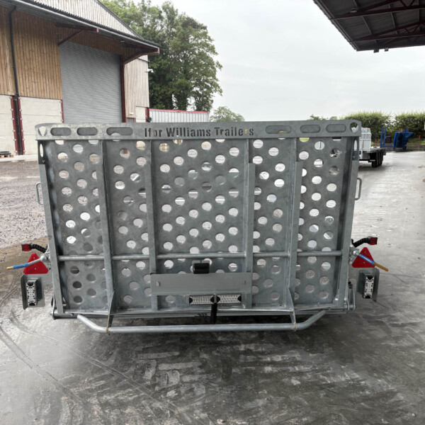Rear view of a gray perforated metal trailer with circular cutouts, parked in a loading yard beside a wooden storage building