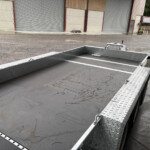 Industrial loading area with a gray flatbed trailer and metal diamond-plate ramp beside closed storage roll-up doors on a wet gravel surface.