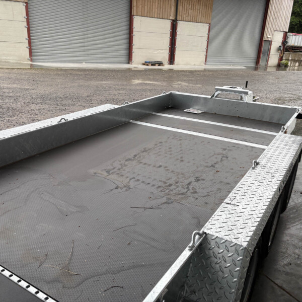 Industrial loading area with a gray flatbed trailer and metal diamond-plate ramp beside closed storage roll-up doors on a wet gravel surface.