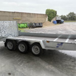 Silver heavy-duty trailer with perforated metal ramps and multiple wheels in an industrial yard beside a concrete wall.