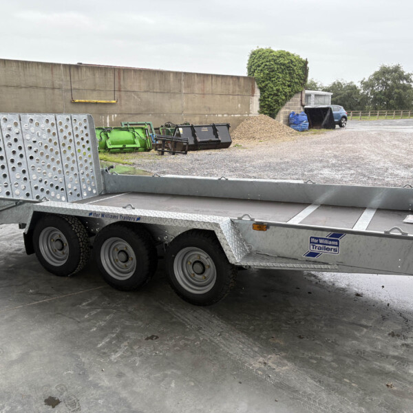 Silver heavy-duty trailer with perforated metal ramps and multiple wheels in an industrial yard beside a concrete wall.
