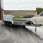 Industrial yard scene with a metal flatbed trailer in the foreground, a concrete wall, wood-chip pile, and a blue car in the background.