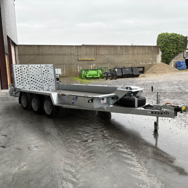 Industrial yard scene with a metal flatbed trailer in the foreground, a concrete wall, wood-chip pile, and a blue car in the background.
