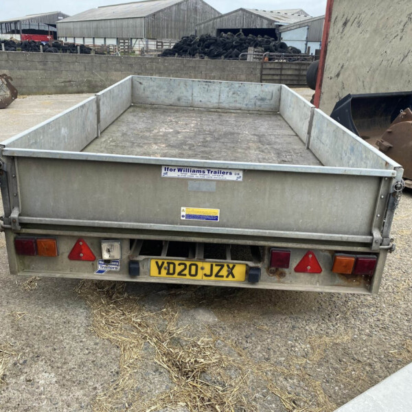 Open-top gray trailer with a yellow license plate reading YD20 JZX, parked in a farm yard; background shows stacked tires, metal sheds, and farm equipment.