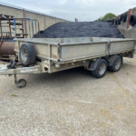 Flatbed trailer loaded with a large pile of dark gravel in an outdoor yard. The trailer sits between concrete walls with tires attached along the side.