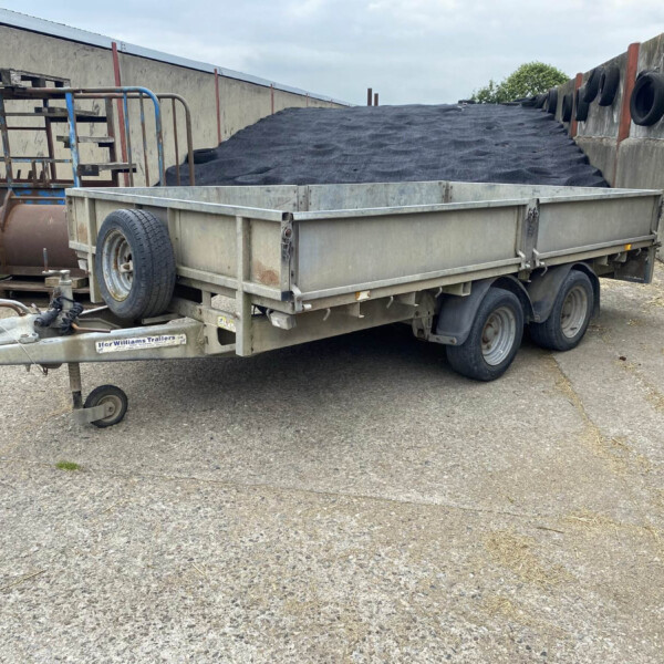 Flatbed trailer loaded with a large pile of dark gravel in an outdoor yard. The trailer sits between concrete walls with tires attached along the side.