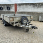Empty metal utility trailer parked in a gravel lot with a spare wheel and hitch in the foreground, against a weathered concrete wall with tires mounted on it behind.