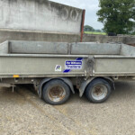 Side view of a weathered metal trailer with rusted wheels, parked on a gravel surface; sticker reads 'For Williams Trailers' with a rural field in the background.