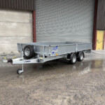 Metal-framed trailer parked in front of a large corrugated shutter and yellow door in an industrial building, wet ground with puddles nearby.