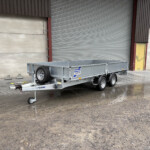 Silver dual-axle utility trailer parked in an industrial yard in front of a large corrugated metal roll-up door; the ground is wet and muddy.
