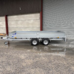 Silver utility trailer parked in front of a brown industrial wall with a large metal shutter, on a wet, muddy concrete surface.