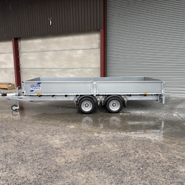 Silver utility trailer parked in front of a brown industrial wall with a large metal shutter, on a wet, muddy concrete surface.