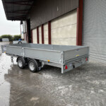 Silver flatbed trailer parked in front of a large industrial warehouse with a corrugated metal facade and a closed roll-up door; wet, muddy ground around it.