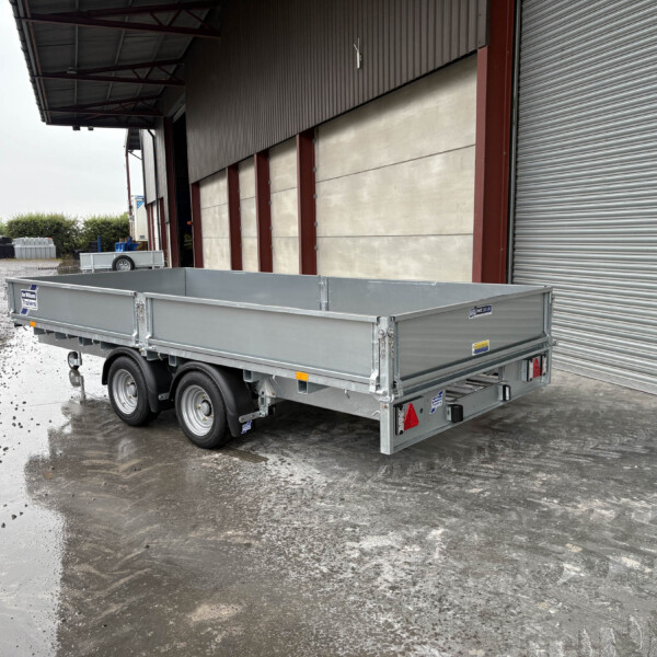 Silver flatbed trailer parked in front of a large industrial warehouse with a corrugated metal facade and a closed roll-up door; wet, muddy ground around it.