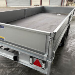 A gray metal flatbed trailer with tall side walls and chains at the rear, parked on a wet concrete surface near storage units.