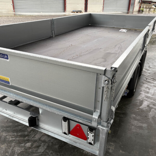 A gray metal flatbed trailer with tall side walls and chains at the rear, parked on a wet concrete surface near storage units.