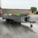 Grey work trailer hitched to a small truck in an industrial yard with gravel piles and a blue car in the background.