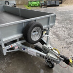 Close-up of a gray metal utility trailer with a mounted spare tire, hitch, and hydraulic system, parked on a wet gravel yard.