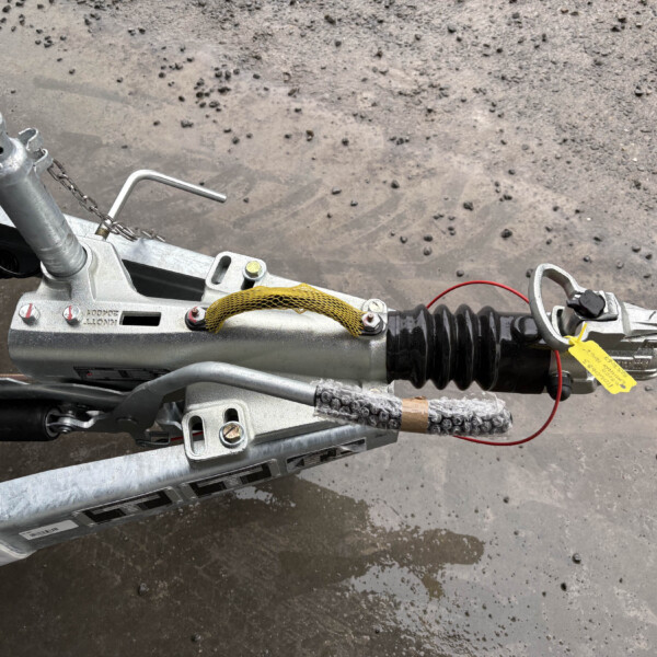Close-up of a trailer hitch tongue with jack, safety chain, and yellow safety strap on a muddy asphalt surface.
