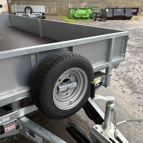 Gray metal utility trailer with a mounted spare tire on the side, in a gravel lot with dumpsters and equipment in the background understood as an outdoor storage area.