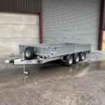 Silver metal utility trailer with dual axles and hitch, parked in front of a corrugated metal wall and large gray roller shutter.