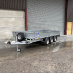 Industrial yard with a metal utility trailer parked near a closed corrugated metal door; trailer has a spare wheel and hitch visible on wet ground.