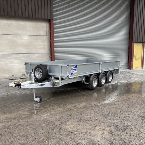 Industrial yard with a metal utility trailer parked near a closed corrugated metal door; trailer has a spare wheel and hitch visible on wet ground.