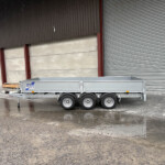 A metal trailer parked on a wet concrete surface in front of a large industrial building with a corrugated metal wall and closed roller door; logo reads 'Ifor Williams Trailers' on the side panel.