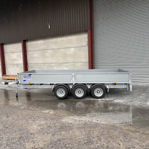 A metal trailer parked on a wet concrete surface in front of a large industrial building with a corrugated metal wall and closed roller door; logo reads 'Ifor Williams Trailers' on the side panel.