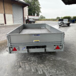 Large gray open-top trailer viewed from behind in an industrial yard with a car and a truck trailer in the background.