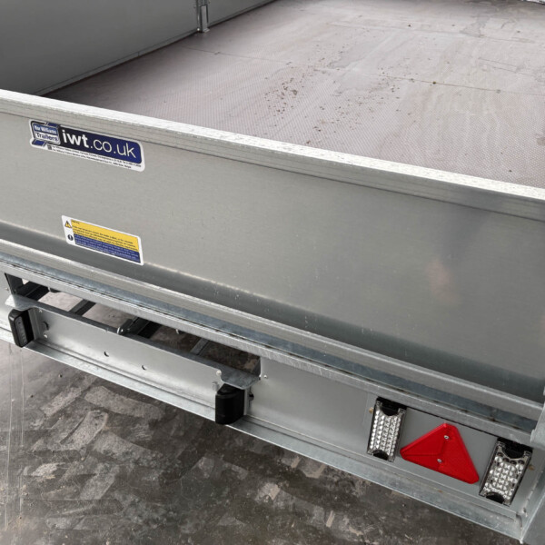 Rear corner of a metal trailer bed with a blue iwt.co.uk sticker and warning labels, showing a diamond-plate floor and red reflectors on the side.