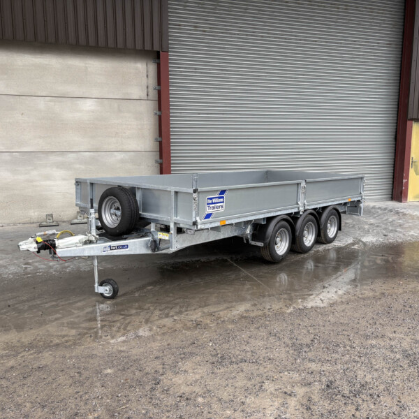 Silver metal utility trailer with dual axles and hitch, parked in front of a corrugated metal wall and large gray roller shutter.