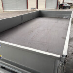 Empty grey metal flatbed trailer with textured floor, outdoors near storage units and a black car in the background