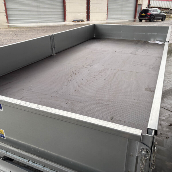 Empty grey metal flatbed trailer with textured floor, outdoors near storage units and a black car in the background