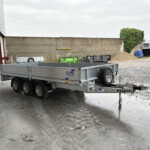Industrial outdoor yard with a silver trailer in the foreground, a tall concrete wall behind, and vehicles and piles of gravel in the background.