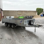 Flatbed trailer with spare tire parked outside a warehouse, beside a gray corrugated door and concrete wall.