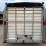 Rear view of a metal trailer in a gravel lot, showing closed tailgate, red taillights, and bottom red reflective triangles; a blue sticker is on the tailgate.