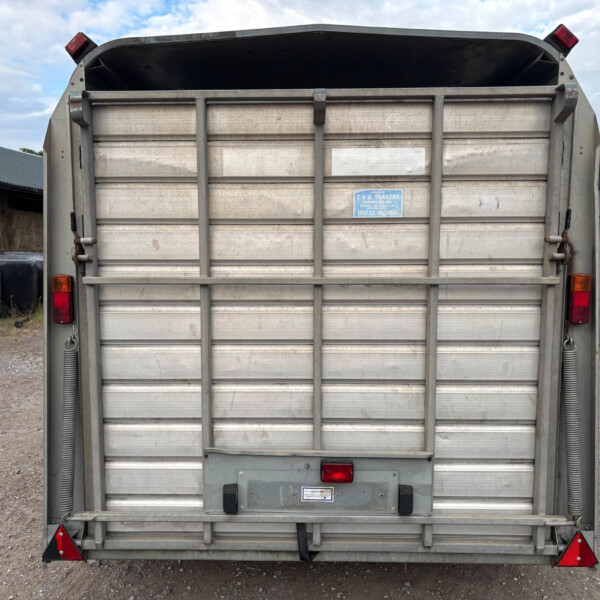 Rear view of a metal trailer in a gravel lot, showing closed tailgate, red taillights, and bottom red reflective triangles; a blue sticker is on the tailgate.