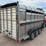 Metal livestock trailer hitched to a red pickup in a farmyard under a partly cloudy sky.